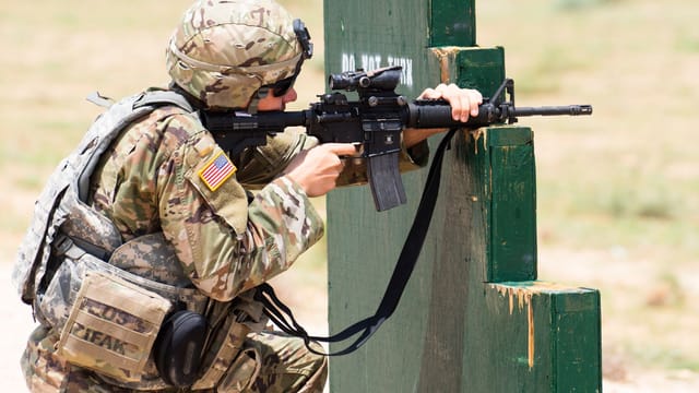 Soldier in camouflage takes aim with a rifle during training, in an outdoor setting.