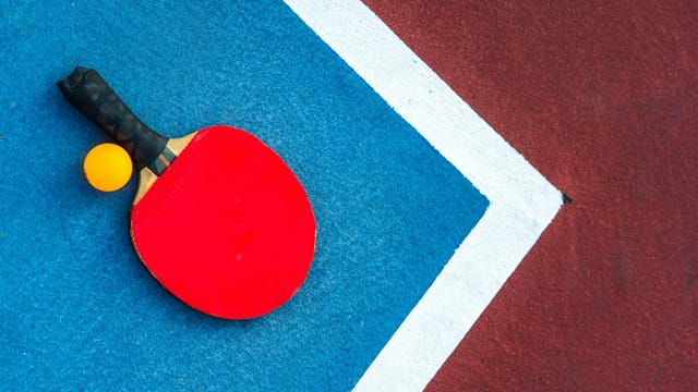 Red table tennis paddle and orange ball on vibrant blue and red court with white lines.