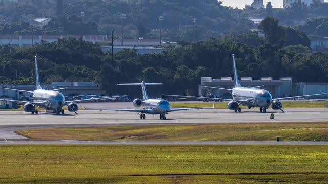 Multiple commercial airplanes on taxiway at airport surrounded by lush greenery under clear daylight.