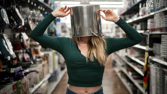A playful scene of a woman with a pot on her head in a store's kitchenware aisle.