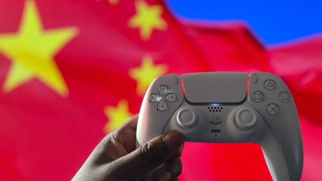 Close-up of a hand holding a PlayStation controller in front of a Chinese flag. Captured in London, UK.