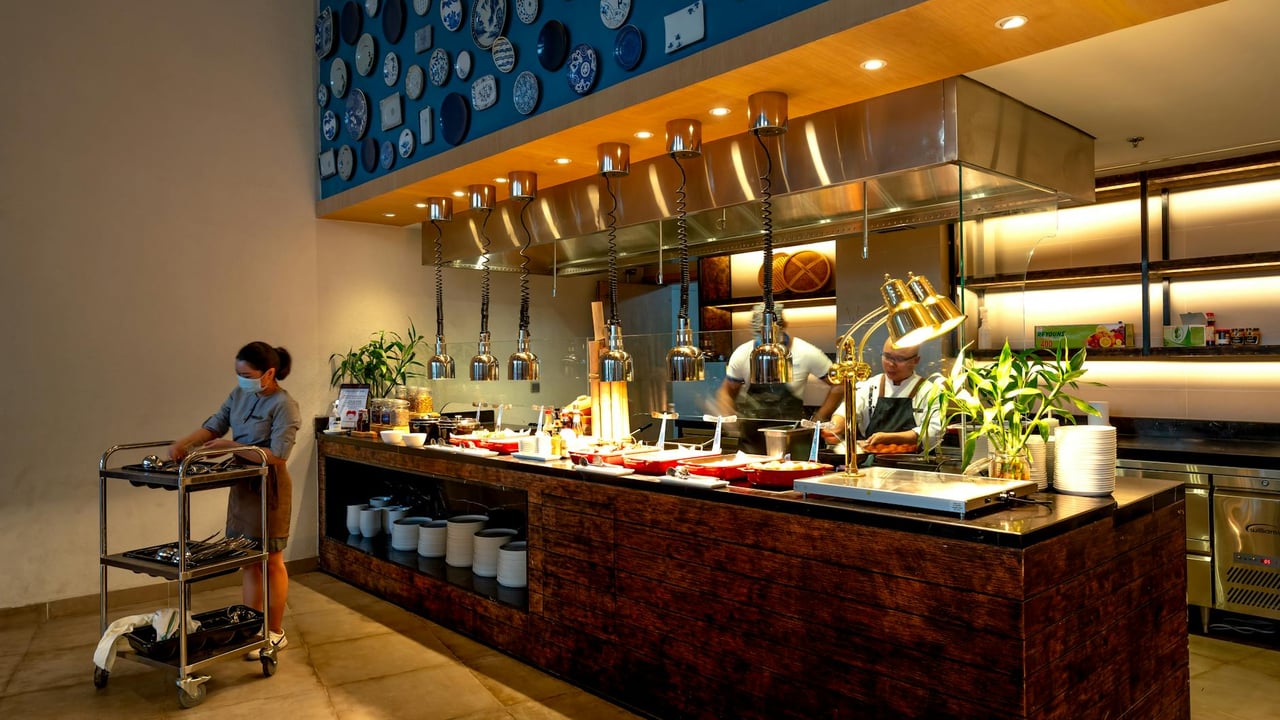 Modern restaurant kitchen with chefs preparing food and waitress on side.