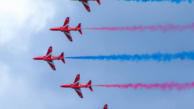 Five Red Arrows jets performing a vibrant aerobatic display against a clear sky.
