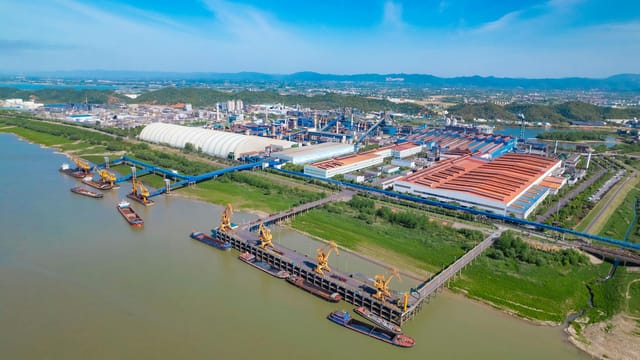 A spacious industrial port with yellow cranes, warehouses, and river access under a clear blue sky.