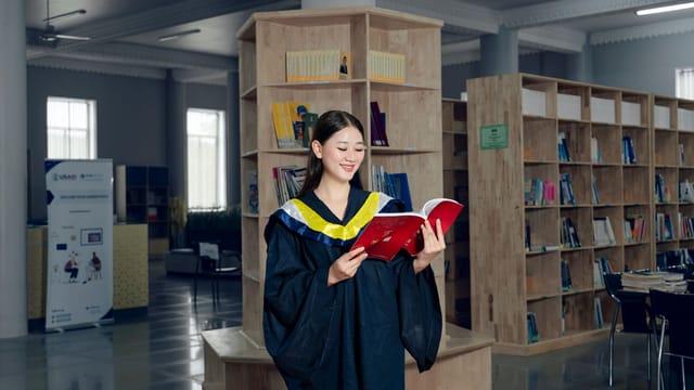 Woman in graduation gown reading a book in university library, symbolizing education and knowledge.