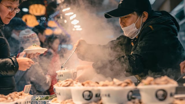 Bustling street food scene in Beijing with vendor serving steamed dishes to customers in the night market.