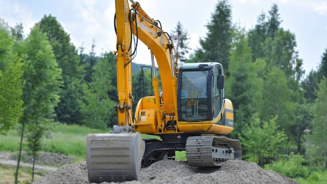 A yellow excavator on a pile of gravel in a lush, green forest setting in Poland.