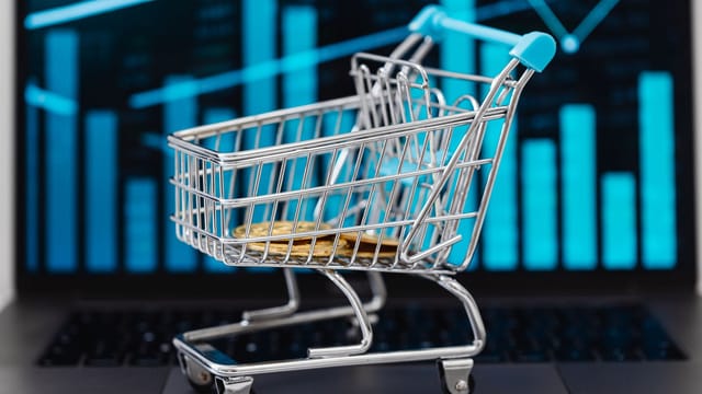 A miniature shopping cart with coins sits on a laptop displaying financial graphs, symbolizing e-commerce and online shopping trends.