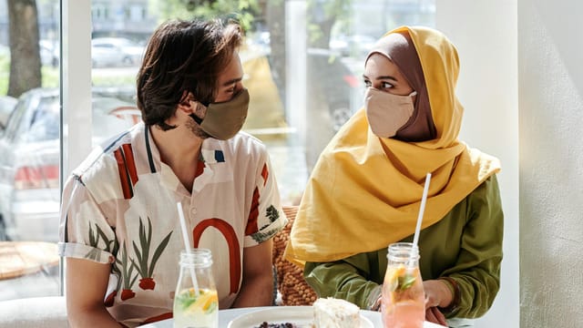 A young couple in masks sharing a meal in a cozy cafe, embracing togetherness.