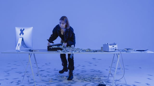 A young woman standing at a desk filled with money, suggesting cyber crime or financial scam.