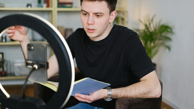 Adult male recording video content indoors, using a smartphone and ring light, holding a notebook.