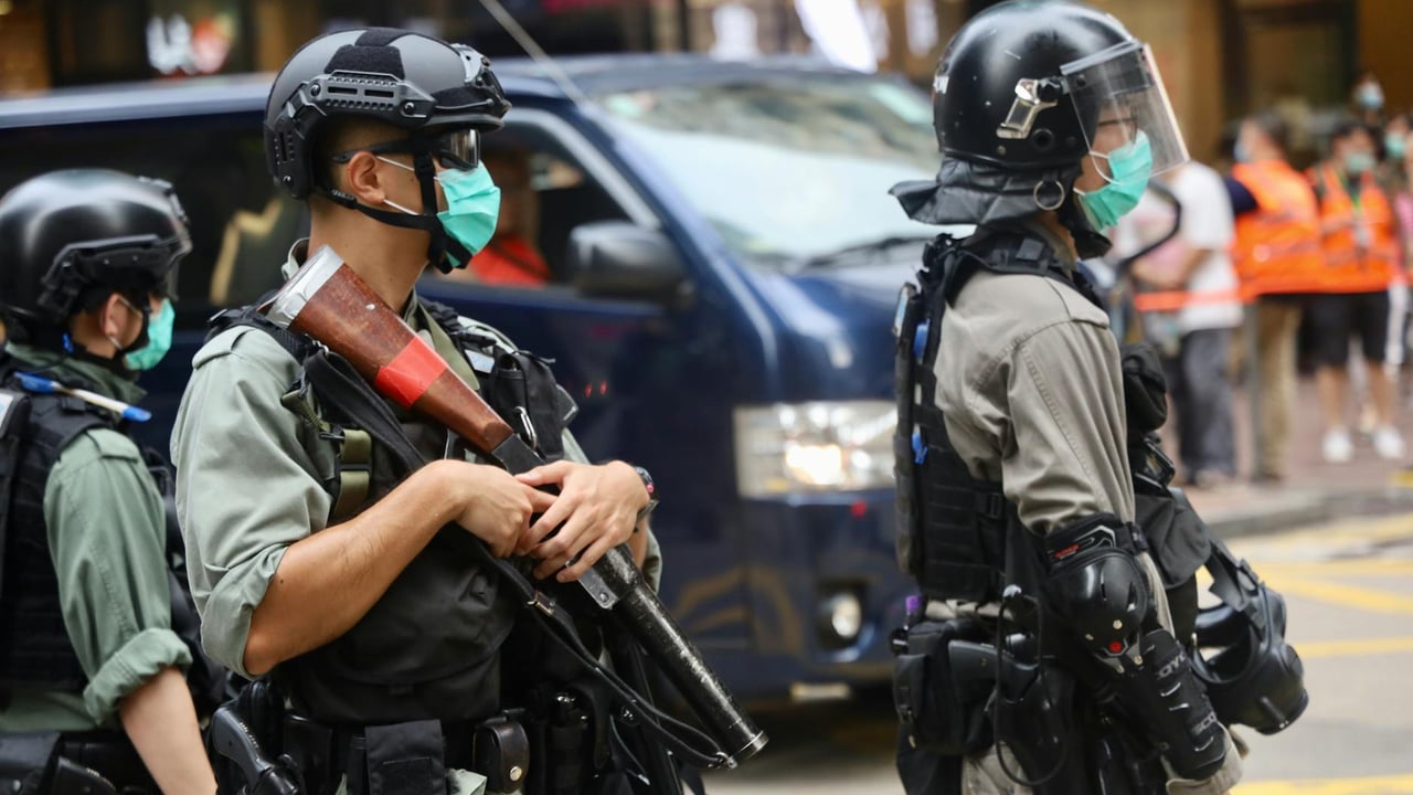 Police officers wearing riot gear and face masks stand in an urban street with protective equipment.