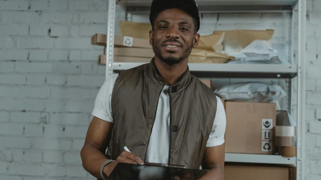 Warehouse employee holding a clipboard, surrounded by packages and smiling.