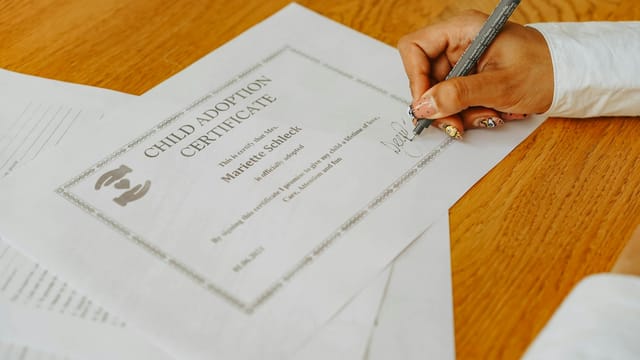 A close-up of a hand signing a child adoption certificate with a pen on a wooden table.