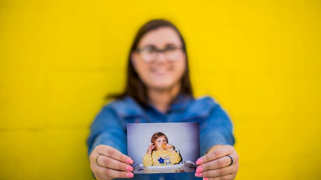 A smiling woman holding a nostalgic childhood photo against a vibrant yellow backdrop.