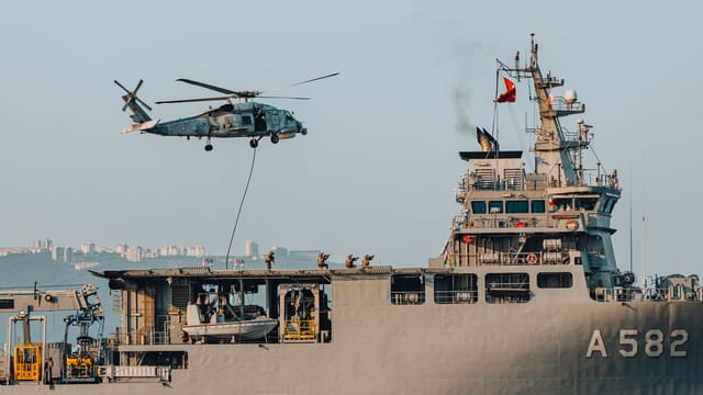 A military helicopter performs an operation above a naval ship at sea, showcasing precision and teamwork.