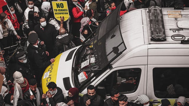 A crowd of protesters surrounding a police van holding signs during a demonstration against Israeli occupation.