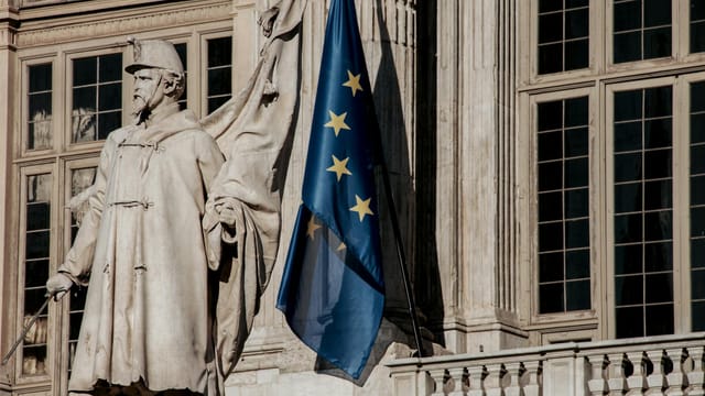 European Union flag waving beside a historic statue on stone building facade.