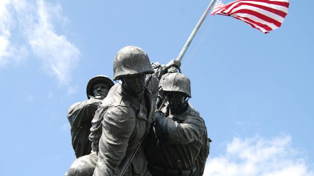 Dramatic view of the Iwo Jima Memorial under a blue sky, featuring US Marines raising the American flag.