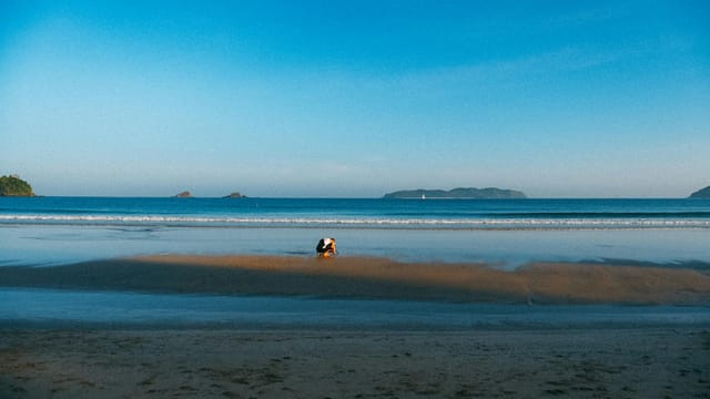 A person squats on a tranquil sandy beach with clear sky and calm sea in the Philippines.