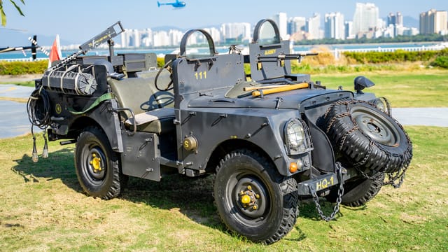 A classic army jeep displayed on grass with a helicopter and skyline in the background.
