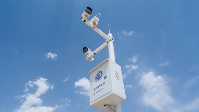 White security cameras mounted on a pole against a bright blue sky with fluffy clouds.