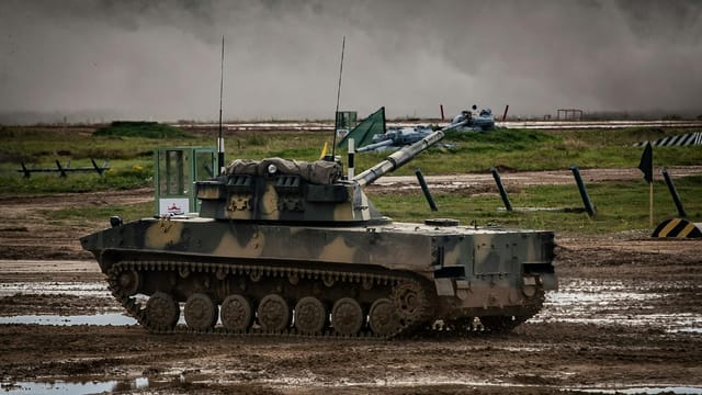 A military tank moves through a muddy training ground with dust in the background.