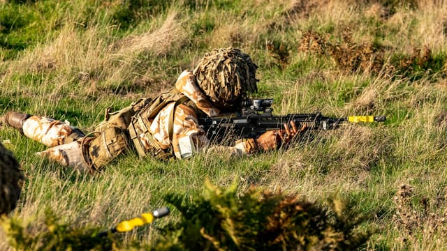 Soldier in camouflage gear practicing tactical maneuvers in a grassy field in Thetford, UK.