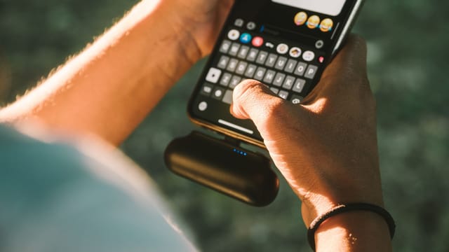 Illustrative image: Close-up of a person texting on a smartphone connected to a power bank outdoors.