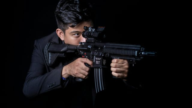 Portrait of a man aiming a rifle with a focused expression against a black background.