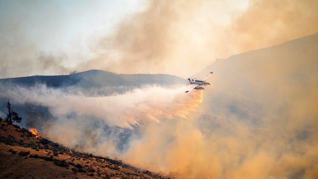 A firefighting plane combats a wildfire, spreading water over rugged terrain at sunset.