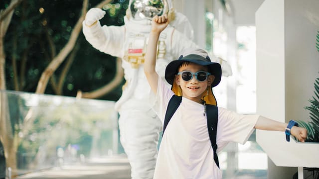 A smiling child in a hat and sunglasses posing beside an astronaut statue in an outdoor setting.
