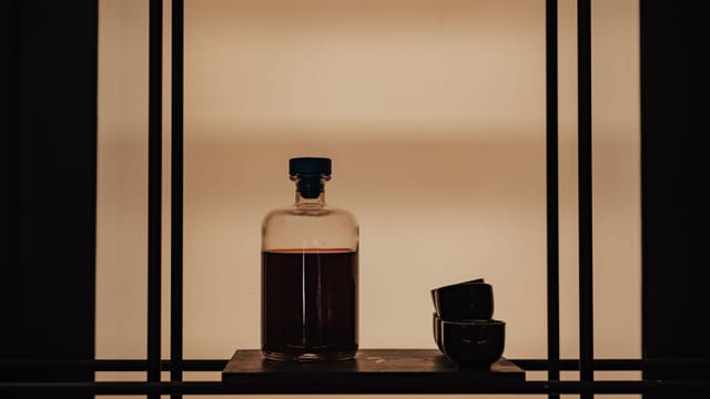 Moody still life featuring a bottle and cups in silhouette on a minimalist shelf.