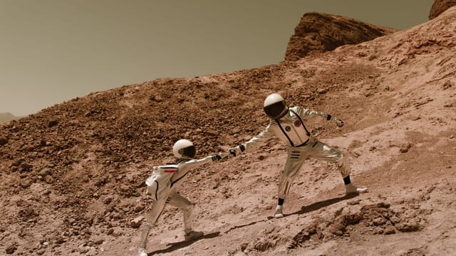 Two astronauts in space suits assisting each other on a rocky terrain resembling Mars.