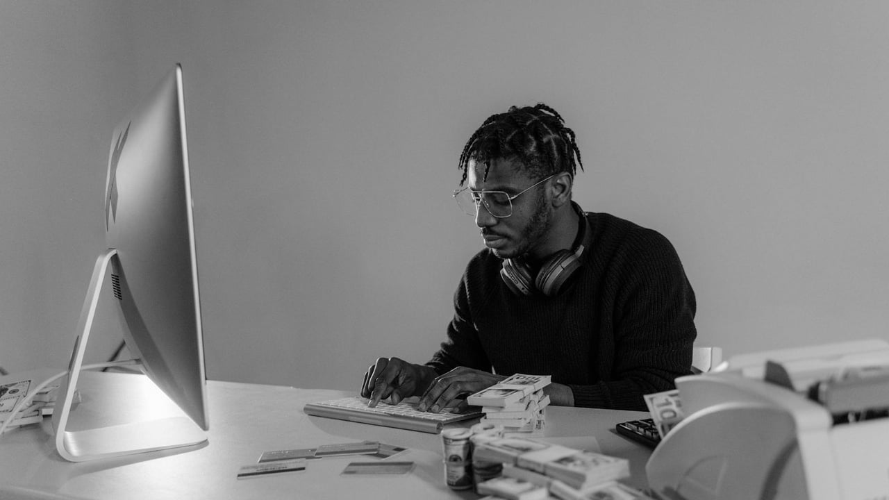 Black and white photo of a focused man typing surrounded by cash indoors.