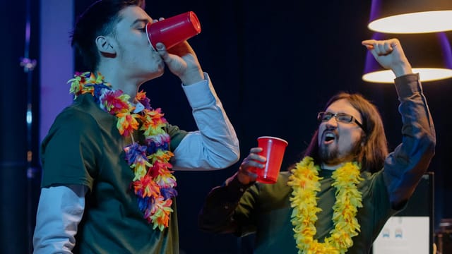 Two men celebrating at an office party with leis and red cups, exuding excitement.