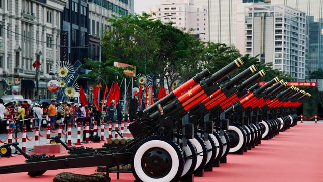 Line of military cannons in an urban area with buildings in the background.