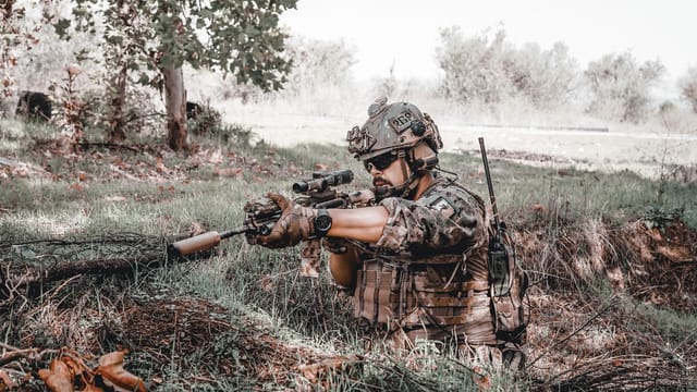 Military personnel in action, wearing camouflage and aiming a rifle outdoors.