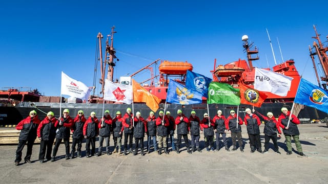 A group of explorers with flags in front of an icebreaker on a sunny day.