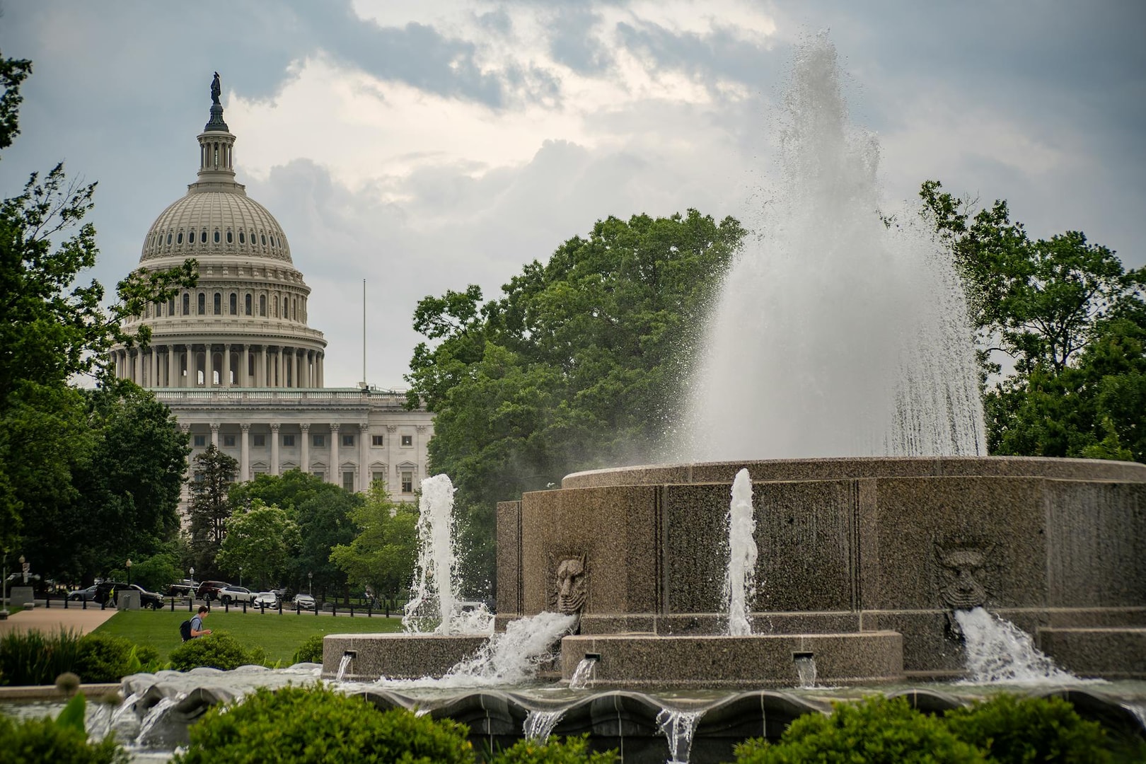 A scenic view of the US Capitol building with a fountain in the forefront, surrounded by lush greenery.