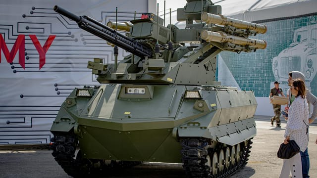 Green military tank with missile launchers on display outdoors with spectators.