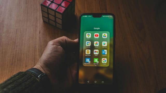 A hand holds a smartphone displaying various apps, next to a Rubik's cube on a wooden surface.