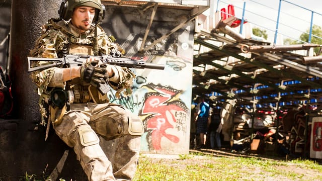 Young soldier in camouflage gear resting against a pillar with a rifle. Urban background.