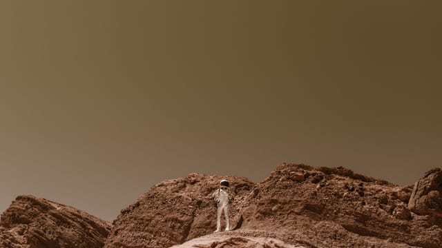 Astronaut in a silver space suit standing on barren rocky terrain, resembling Mars.