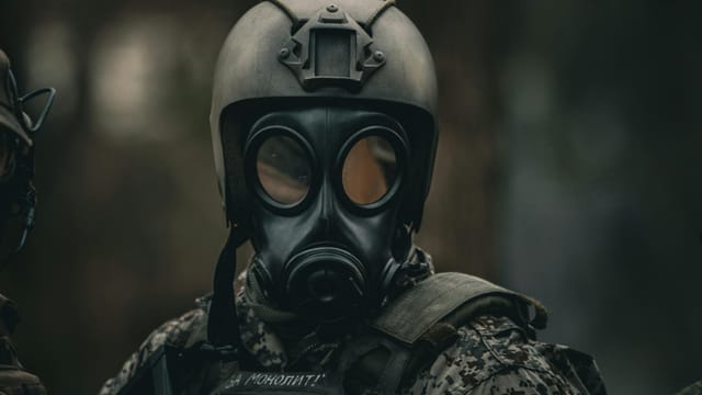 A soldier wearing a gas mask and camouflage gear stands in a dense forest.
