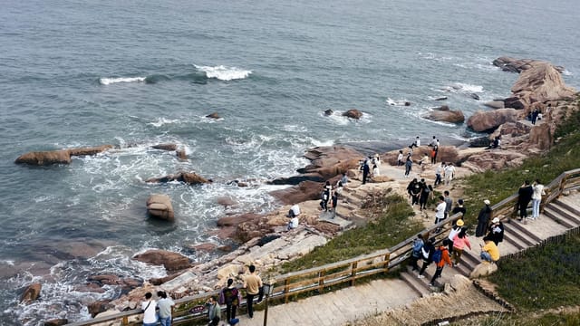 From above of distant unrecognizable travelers walking on stony coast and admiring view of sea with waves crashing against rocks