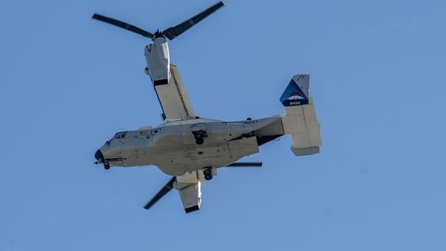 Tiltrotor military aircraft flying against blue sky, seen from below.