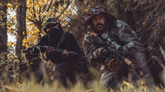 Two soldiers in camouflage gear patrol a forested area, ready for combat.