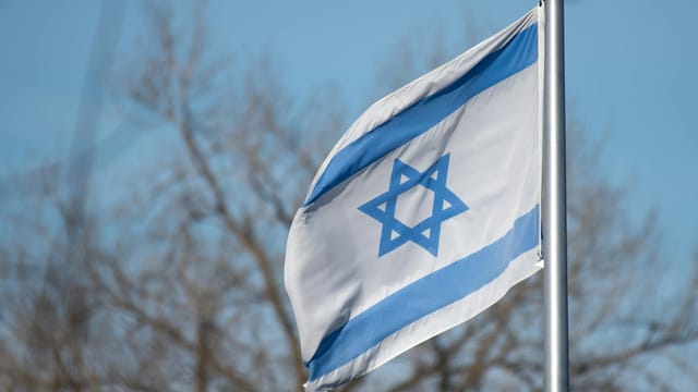 The Israeli flag featuring the Star of David against a clear blue sky and trees.