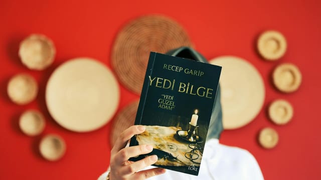 Person holding 'Yedi Bilge' book in front of a red wall with decorative plates. Close-up focus on book.